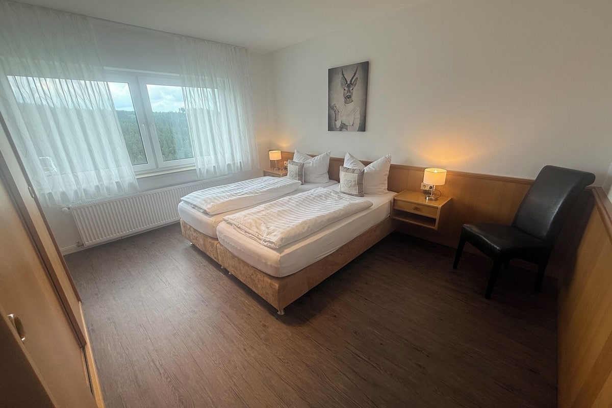 A hotel room with a double bed, two bedside tables with lamps, a chair, a window with sheer curtains, and a painting on the wall.
