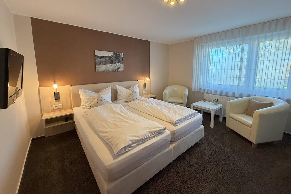 A hotel room with a large bed, two bedside tables, a chair, a small table, and a window with sheer curtains.