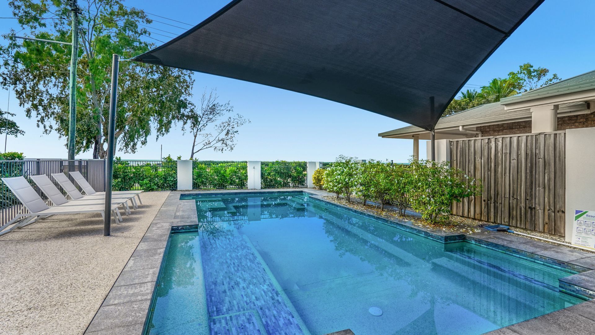 Piscina rectangular con un toldo oscuro, rodeada por una cerca de madera, un suelo de hormigón y pequeños árboles.