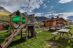 Explore the great outdoors at this scenic play area, featuring a rustic play structure with a slide and climbing area, surrounded by picnic tables and a serene natural setting.