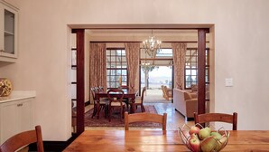 A dining area with a wooden table, chairs, and a fruit bowl. There is a dining table set with chairs, a chandelier, and a view of a living room through an open doorway. - Dog-friendly Farm Cottage in the Berg by First Private Stays (Drakensberg Gardens)