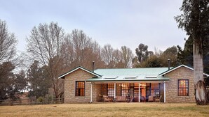 A stone house with a green roof, a porch with chairs, and a fenced yard. - Dog-friendly Farm Cottage in the Berg by First Private Stays (Drakensberg Gardens)