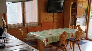 A wooden dining area with a table covered in a floral tablecloth, wooden chairs, a television mounted on the wall, and a window with lace curtains. - Cozy Apartment for 4-6 in Village Center with Terrace and Parking (BLA003) (Saint-Jean-de-Sixt)