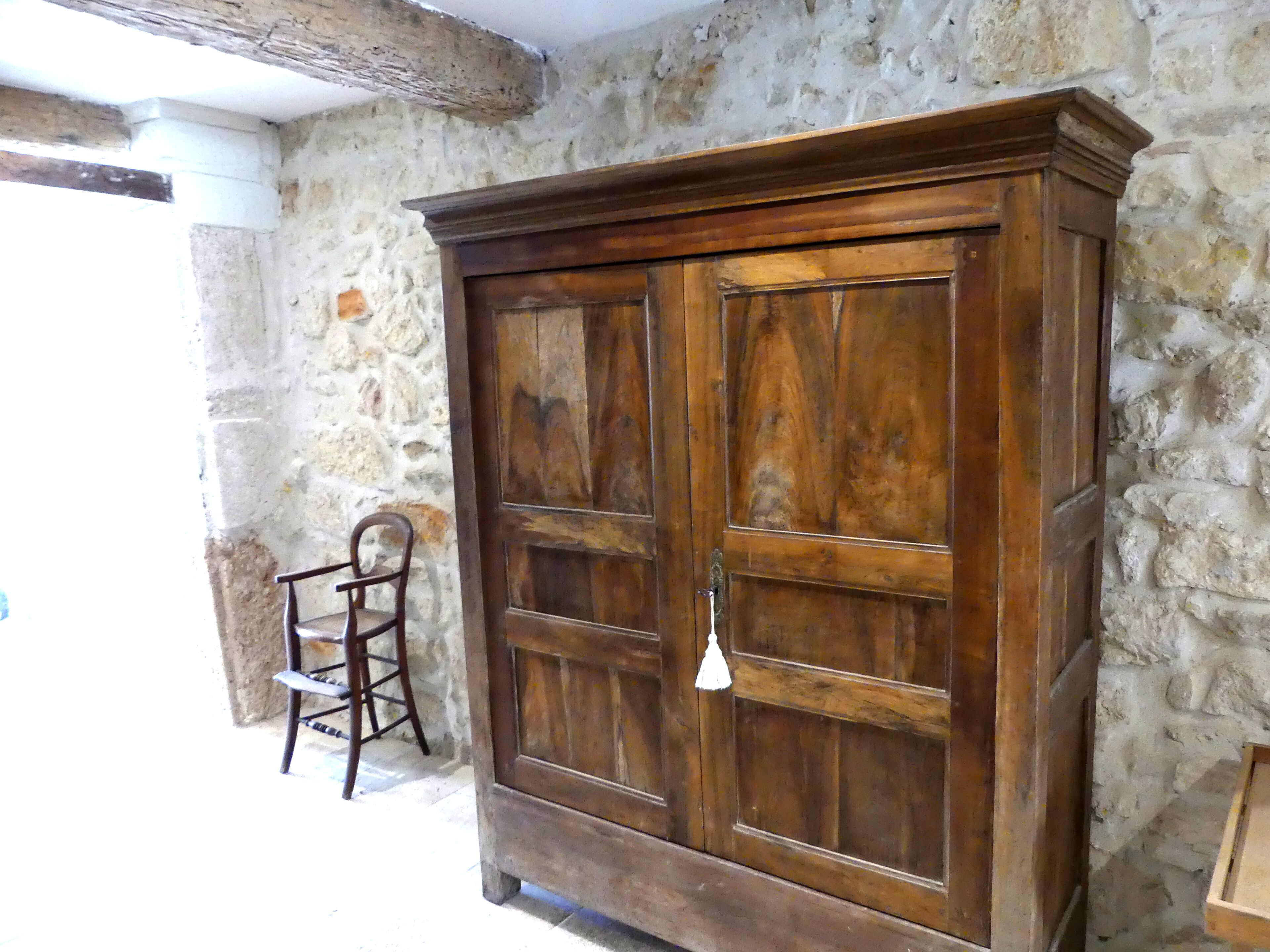 A wooden wardrobe with a large mirror and a chair in a stone-walled room.