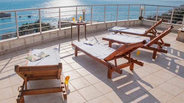 A terrace with wooden lounge chairs and a table, overlooking a body of water and distant hills.