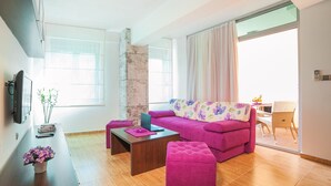 A modern living room with a pink sofa, a wooden coffee table, and a small ottoman. There is a TV mounted on the wall, a potted plant, and a window with curtains.