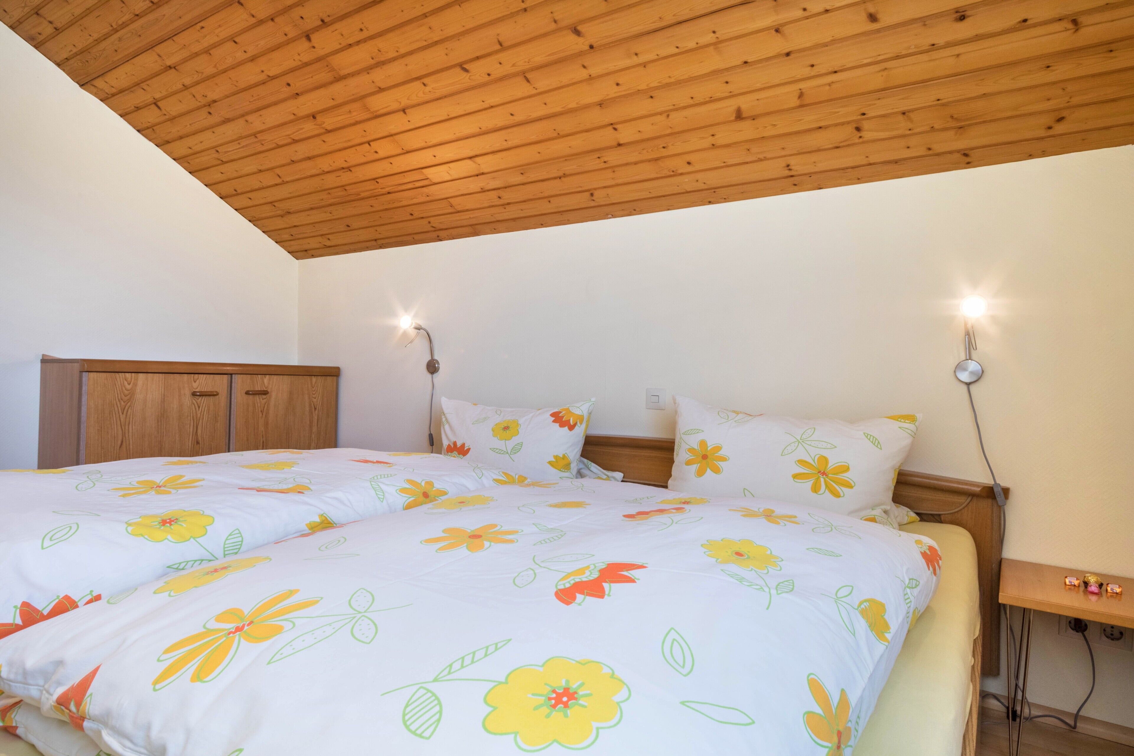 Une chambre avec un plafond en bois, un lit recouvert d’une literie à motifs floraux, des lampes de chevet et une table de chevet en bois.