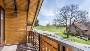 A wooden balcony with a view of a rural landscape, including a tree and a building in the distance.