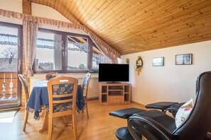 A cozy living room with a wooden ceiling, a television on a wooden stand, a dining table with chairs, and a black leather sofa.