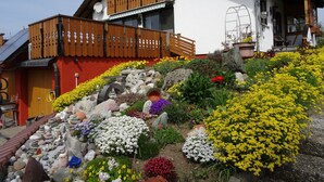 A house with a wooden balcony, a garden with various plants and flowers, and a stone pathway.