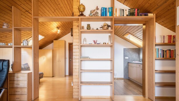 A spacious loft with wooden beams, a mezzanine with bookshelves, and a kitchen area.