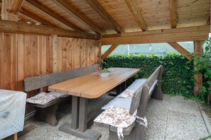 A wooden outdoor dining area with benches and a table, surrounded by wooden walls and a hedge.