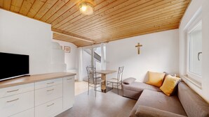 Un intérieur moderne avec un plafond en bois, un téléviseur à écran plat, un coin repas avec des chaises, un canapé avec des coussins et une croix accrochée au mur.