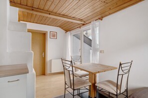 Ein Zimmer mit Holzbalkendecke, einer weißen Tür, einem Holztisch mit Stühlen und einem Fenster mit Vorhängen.