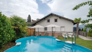 Une piscine avec une terrasse en bois, une maison à la façade blanche et une cour clôturée.
