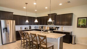 A modern kitchen with dark cabinets, a stainless steel refrigerator, a large island with bar stools, and pendant lighting.