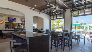 A spacious outdoor bar area with high tables and chairs, a counter with a bar, and a view of a pool and palm trees.