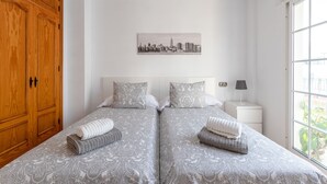 Une chambre d’hôtel avec deux lits, une armoire en bois, une œuvre d’art représentant un paysage urbain, une lampe de chevet et une fenêtre avec vue.