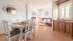 Un coin repas avec une table ronde et des chaises, un canapé, une cuisine avec des meubles en bois et une horloge murale.