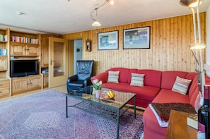 A cozy living room with a red sofa, a black leather chair, a glass coffee table, wooden bookshelves, and a television.