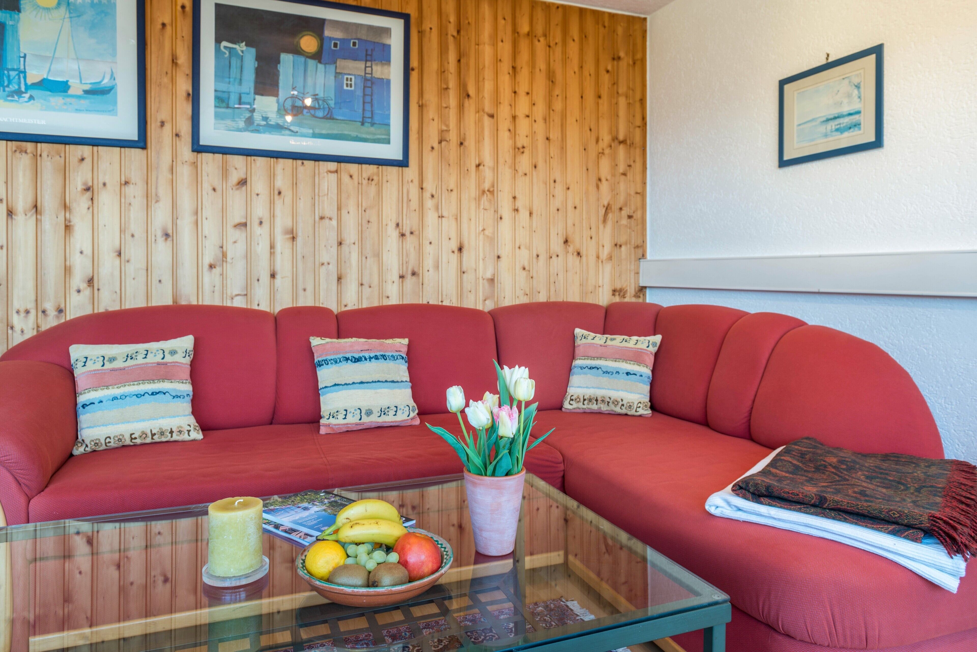 Ein rotes Sofa mit gemusterten Kissen, ein Glastisch mit Obst und einer Kerze sowie Holzvertäfelung an der Wand.