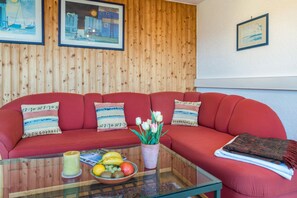 A red sofa with patterned cushions, a glass coffee table with fruit and a candle, and wooden paneling on the wall.