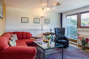 A living room with a red sofa, a black leather chair, a glass coffee table, and a view of greenery outside.