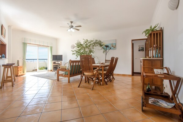 A spacious living area with a dining table, chairs, a ceiling fan, a large window, and a view of the sea.