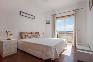A bedroom with a large bed, a nightstand, a dresser, a window with curtains, and a balcony with a view.