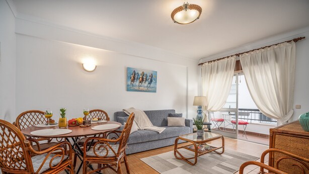 Een woonkamer met een eettafel, rieten stoelen, een bank en een balkon met gordijnen.