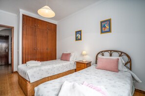 A bedroom with two beds, a wardrobe, a nightstand, and a wall-mounted lamp.