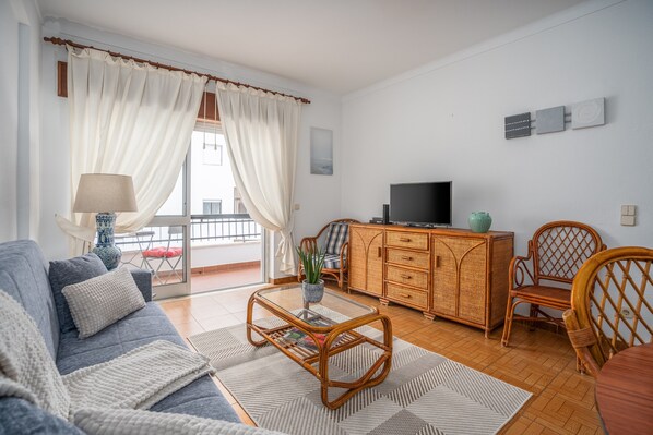 A living room with a sofa, a glass coffee table, wicker furniture, a TV, and a view through a window.