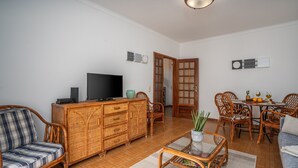 Een woonkamer met een televisie, rieten meubels, een eethoek met een tafel en stoelen, en een deur naar een andere kamer.