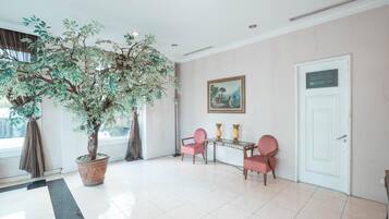 A lobby with a large artificial tree, two chairs, a table with vases, and a door.