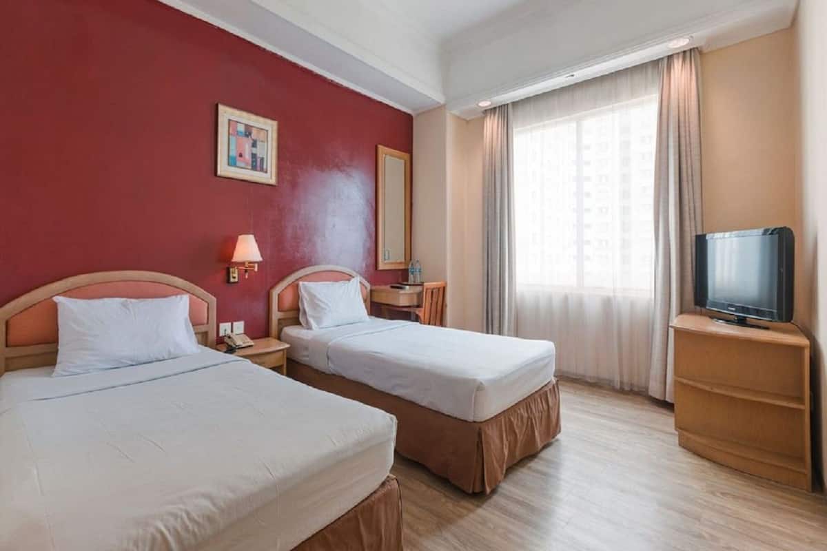 A hotel room with two beds, a TV on a wooden stand, a bedside table with a lamp, and a window with curtains.