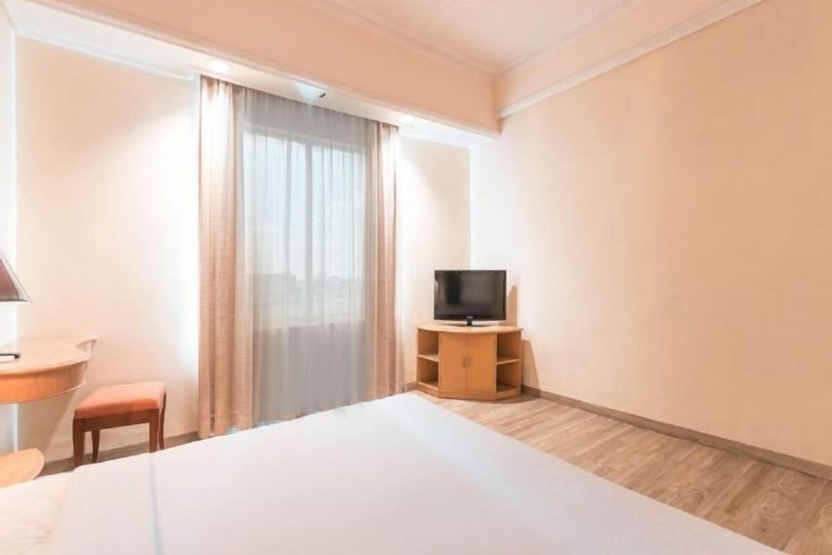 A hotel room with a bed, a TV on a wooden stand, a desk with a chair, and a window with curtains.