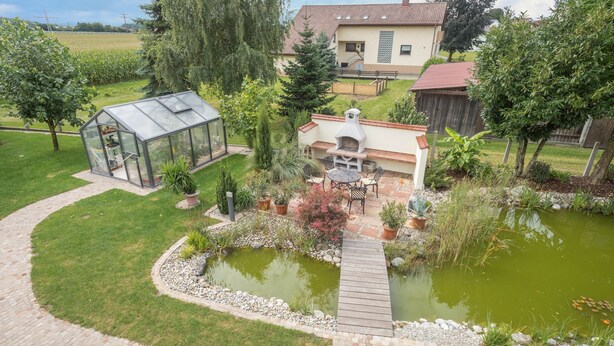 Un jardín residencial con un invernadero, patio, estanque y una casa al fondo.