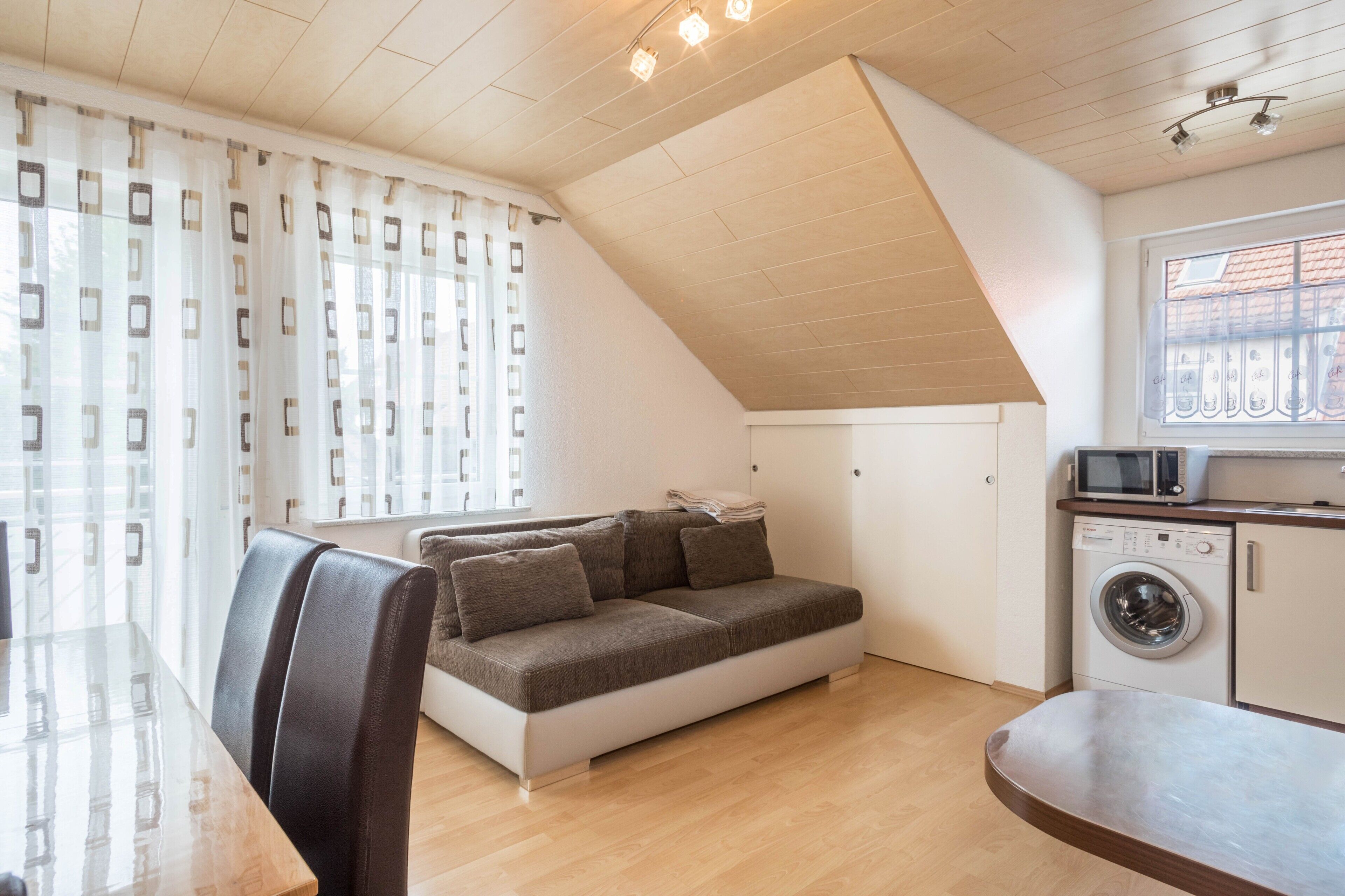 A compact living space with a sofa, a washing machine, a dining table, and a window with curtains.
