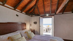 Una camera da letto con soffitto in legno, un letto con cuscini floreali, un comodino e vista sul mare attraverso una porta aperta.