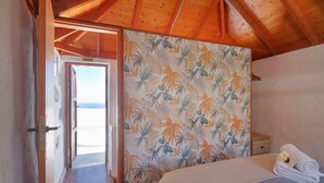 Una camera da letto con soffitto in legno, un letto con copripiumino bianco e una parete decorata con un motivo a foglia tropicale, oltre a una vista attraverso una porta aperta.