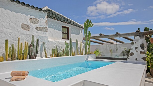 Una piscina con una parete bianca e cactus, un pergolato in legno e una piccola finestra.