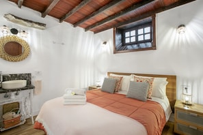 A bedroom with a wooden ceiling, a window, a bed with a pink bedspread, a nightstand with a lamp, and a bathroom with a sink and mirror. - Holiday Home "Casa Pedro" with Terraces, Panoramic Sea View and Wood Stove (Icod de los Vinos)