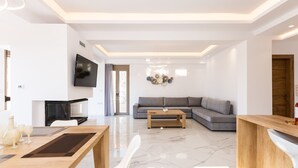 Een moderne woonkamer met een grijze modulaire bank, een houten salontafel, een flatscreen televisie en een eethoek met een houten tafel en witte stoelen.