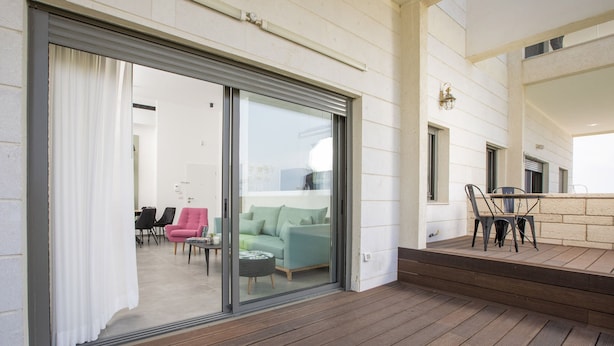 A modern outdoor area with a wooden deck, a sliding glass door leading to a living room with a sofa and chairs, and a small table.