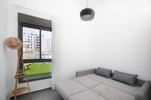 A modern room with a grey sofa, a hanging pendant light, a wooden ladder, and a view of a building and a lawn through a large window.