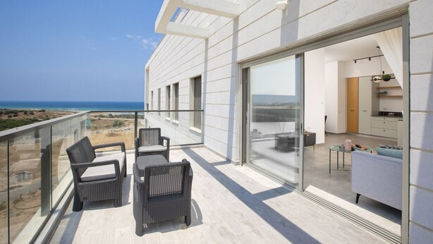 A modern balcony with wicker furniture, a glass door leading to a kitchen, and a view of the coastline. - Chic Sea View Balcony Escape by Sea N' Rent (Nahariya)