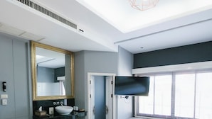 A modern hotel room with a large bed, a bathroom with a sink and mirror, and a window with blinds.