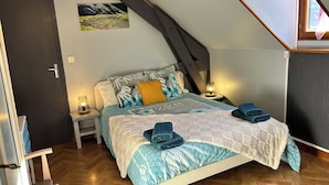A bedroom with a sloped ceiling, a bed with patterned bedding, a nightstand with a lamp, and a painting on the wall. - House with large, quiet garden and beautiful mountain views (ANTIGNAC)