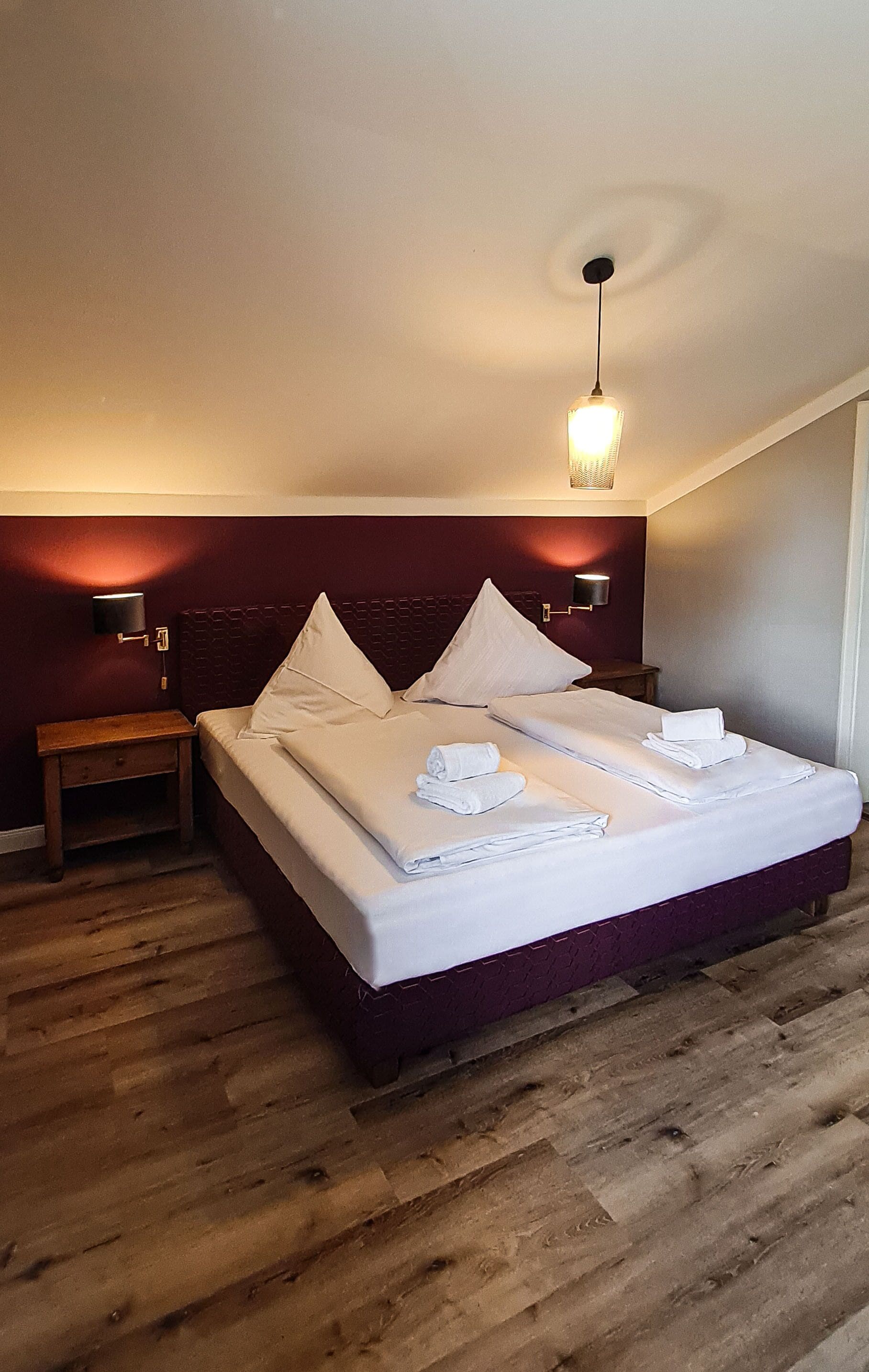 A neatly made bed with white linens and towels, a wooden nightstand, and a wall-mounted light fixture.