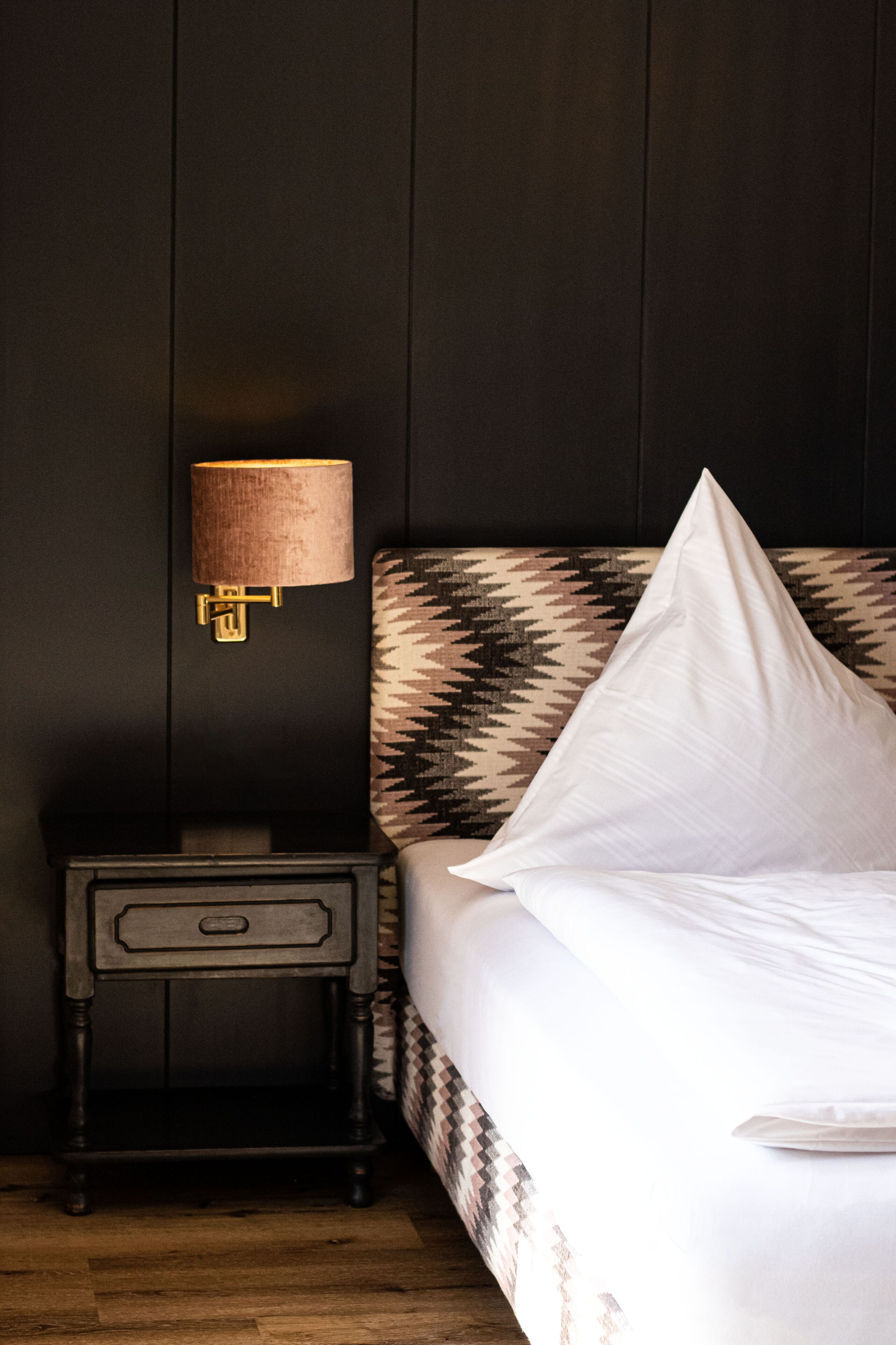 Un lit bien fait, avec une tête de lit à motifs, une lampe de chevet et une petite table de chevet en bois dotée d’un tiroir.